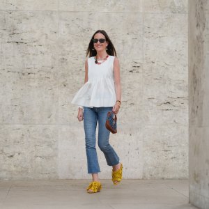 Alba Garavito Torre wears black sunglasses, long straight brown hair with a center part, a white pleated peplum sleeveless blouse by Commense, and blue high-rise raw-hem denim jeans. A yellow clog sandal with floral detailing by AGL is worn, paired with a jean denim and brown mini Jackie handbag by Gucci. Accessories include a chunky necklace with colorful natural stones by Sempiterno Paris and gold jewelry. Full body shot, during a street style fashion photo session, on May 02, 2025 in Paris, France.