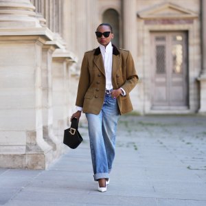 Emilie Joseph wears slicked-back dark brown hair, black sunglasses, a brown khaki vintage military officer jacket by Saint Laurent, a white high-neck shirt, blue wide-leg cuffed jeans with hems, a black bag by Danse Lente white slingback pumps shoes by Saint Laurent, during a street style fashion photo session, on April 24, 2026 in Paris, France.