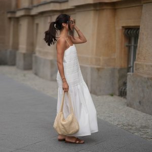 Aylin Sengül is seen wearing a white spaghetti-strap maxi dress with textured horizontal panels and a gathered hem by COS; brown leather “Oran” sandals by Hermès; a raffia shoulder bag with a slouchy hobo shape by COS; a delicate gold-tone necklace; a rose gold “Knot” bracelet by Celine; a vintage tennis bracelet; a slim silver-tone bangle; a gold ring; slim oval dark tortoiseshell sunglasses by Chanel during Berlin Fashion Week Spring/Summer 2027 on July 4, 2026 in Berlin, Germany.