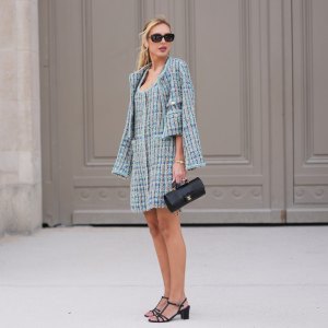 PARIS, FRANCE - JULY 07: A guest wears short straight blonde hair, black rectangular sunglasses with silver chain necklaces, a blue checked pattern tweed jacket from Chanel, a blue checked tweed mini dress from Chanel, a black matte leather shoulder bag from Chanel, black shiny leather straps heels sandals shoes, outside Chanel, during Paris Fashion Week - Haute Couture Fall/Winter 2026/2027, on July 07, 2026 in Paris, France (Photo by Edward Berthelot/Getty Images)