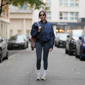 PARIS, FRANCE - OCTOBER 28: Emy Venturini wears straight light brown hair parted in the middle, black rectangular sunglasses by Balenciaga, a navy blue satin bomber jacket by Burberry, a dark grey ribbed top and matching high-waisted leggings by Born Living Yoga, white ribbed socks by Balenciaga, and silver mesh running shoes by Nike, holds a Yoga mat by Decathlon, during a street style fashion photo session, on October 28, 2025 in Paris, France. (Photo by Edward Berthelot/Getty Images)