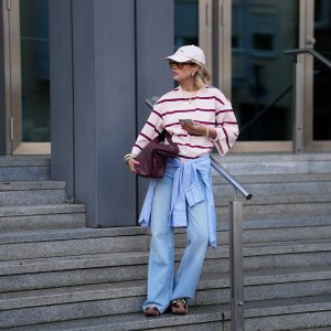 FRANKFURT AM MAIN, GERMANY - MARCH 26: Julia Kammerer is seen wearing an oversized pink and burgundy horizontal striped cotton T shirt from H&M; a blue and white striped button down shirt tied around her waist from JJXX; light blue wide leg denim jeans with a relaxed fit from JJXX; burgundy and yellow textured leather lace up sneakers from Adidas; a large burgundy woven leather hobo bag from NA-KD; a pale pink baseball cap with the red embroidered text "Amore a Milano" from LeGer by Lena Gercke; her jewelry includes chunky gold hoop earrings, necklaces and gold tone stacked rings worn across multiple fingers from H&M, Pandora and My Jewellery; holding an iPhone from Apple; several silver and resin bangles from & Other Stories; orange tinted aviator sunglasses with a thin black frame from Bershka; her blonde hair is styled in loose waves underneath the cap on March 26, 2026 in Frankfurt am Main, Germany. (Photo by Moritz Scholz/Getty Images)