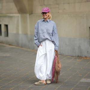 Julia Kammerer is seen wearing a black and white check cotton long sleeved button down shirt from Pull & Bear; white cotton balloon trousers with an elasticated waistband from Bershka; taupe suede „Boston“ clogs with a rounded toe and adjustable strap with a copper tone buckle from Birkenstock; a large tan suede shoulder bag with a curved hobo silhouette, long fringe trim and silver tone stud details from Pull & Bear; rectangular tortoiseshell acetate eyeglasses with clear lenses from & Other Stories; a washed red cotton baseball cap with red embroidered text from New Yorker; a pale pink manicure; several chunky jewelry pieces in gold and silver including rings and hoop earrings from Pandora, Akind, & other stories, H&M, my jewellery or scandiv; her blonde hair is styled in a straight long bob; with a natural make up look on April 21, 2026 in Frankfurt am Main, Germany.