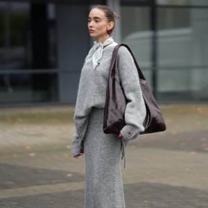 DUESSELDORF, GERMANY - OCTOBER 14: Evi Wave is seen wearing an oversized boatneck grey pullover by Aylin Koenig x Anita Hass with a slouchy fit, dropped shoulders and elongated sleeves made of a soft alpaca mix, it features an embroidered red heart one cuff; a full-length tube grey skirt by Aylin Koenig x Anita Hass in matching soft alpaca mix, with straight silhouette with a slightly flared hemline forming a co-ord set with the top; under the top she layered a white crewneck t-shirt; she carries an oversized shoulder hobo bag by Arket in high-shine patent leather in burgundy with a slouchy body; a pair of pointed-toe beige slingback kitten heels in monogram canvas by Gucci, featuring a brown logo-jacquard pattern, the GG motif with a low stiletto heel and brown leather piping around the edges, these worn with white ankle socks; on her fingers she wears silver rings from Ariane Ernst and Fossil; her nails are polished in red; she wears a silk carré Fraas scarf with a polka-dot print in monochrome black and white tones, tied in a classic French knot at the collar; she wears a pair of sculptural beaded earrings handmade with red glass beads by Kroon02; her brown hair is styled in a sleek, low bun on October 14, 2025 in Duesseldorf, Germany. (Photo by Moritz Scholz/Getty Images)