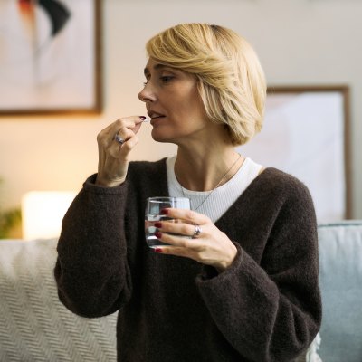 Middle aged Caucasian woman sitting indoors holding glass of water and taking pill, demonstrating self care and health management, focused expression