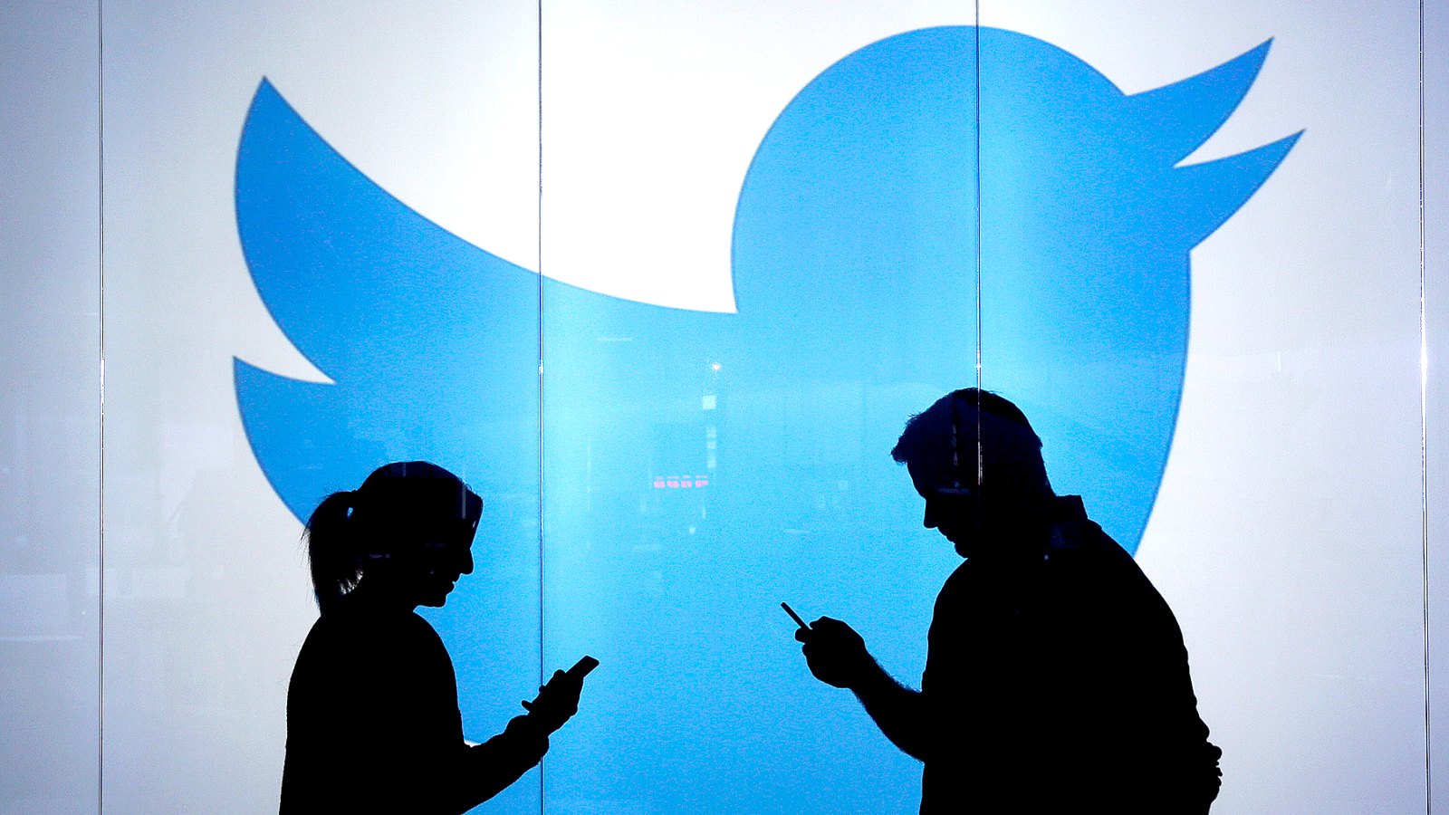 People are seen as silhouettes as they check mobile devices whilst standing against an illuminated wall bearing Twitter Inc.'s logo.