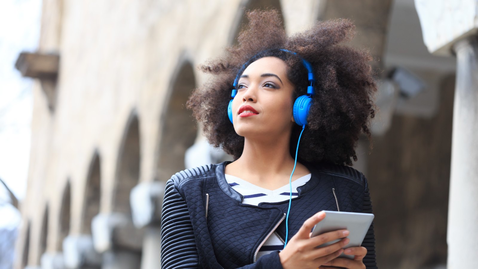 woman with curly afro looking away from the camera while standing outside with blue head phones around her head that are connected to a tablet