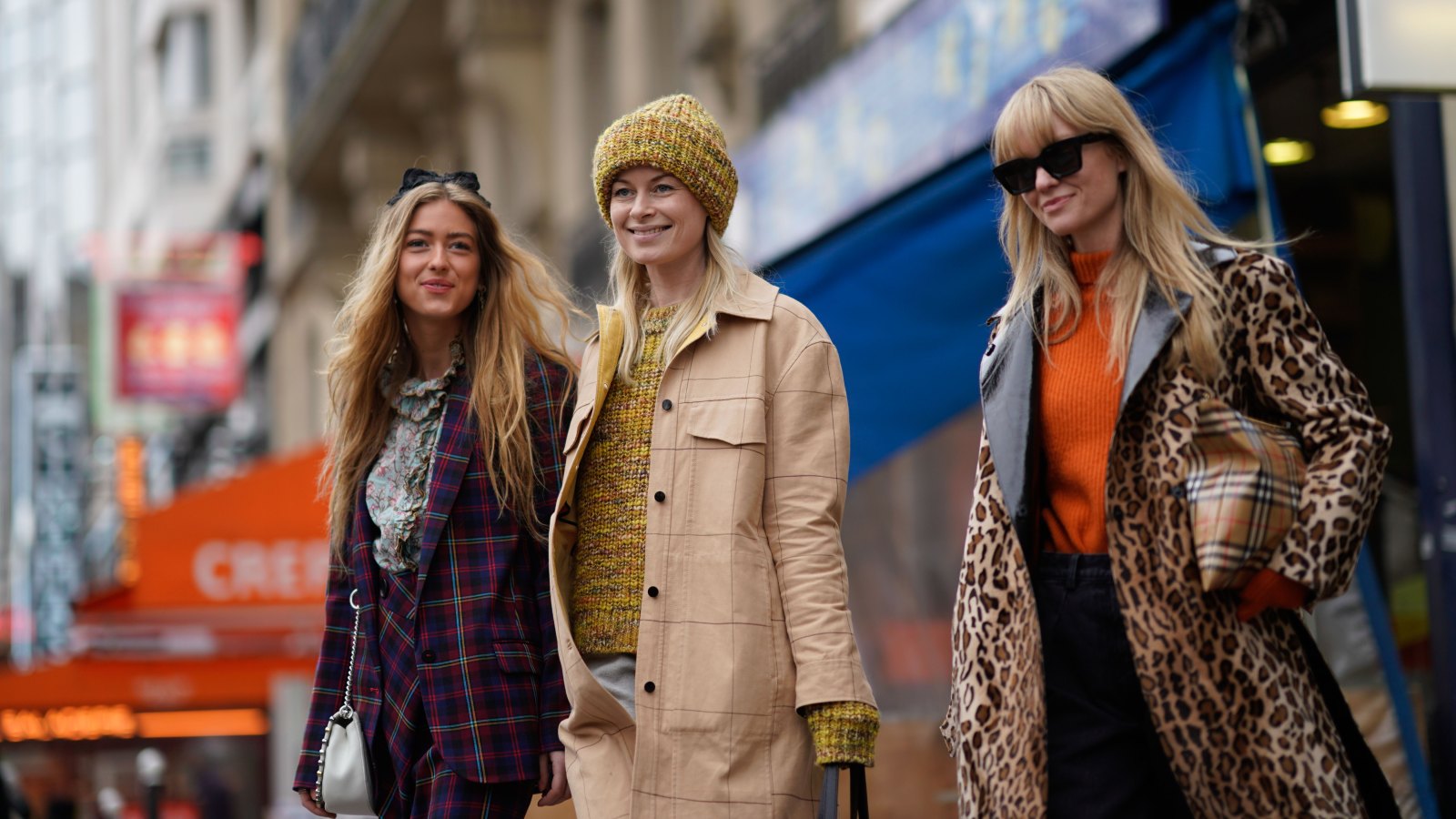three women walking wearng coats. One woman is wearing a khaki coat, another is wearing a plaid blazer coat another is wearing a leopard print coat. All are smiling.