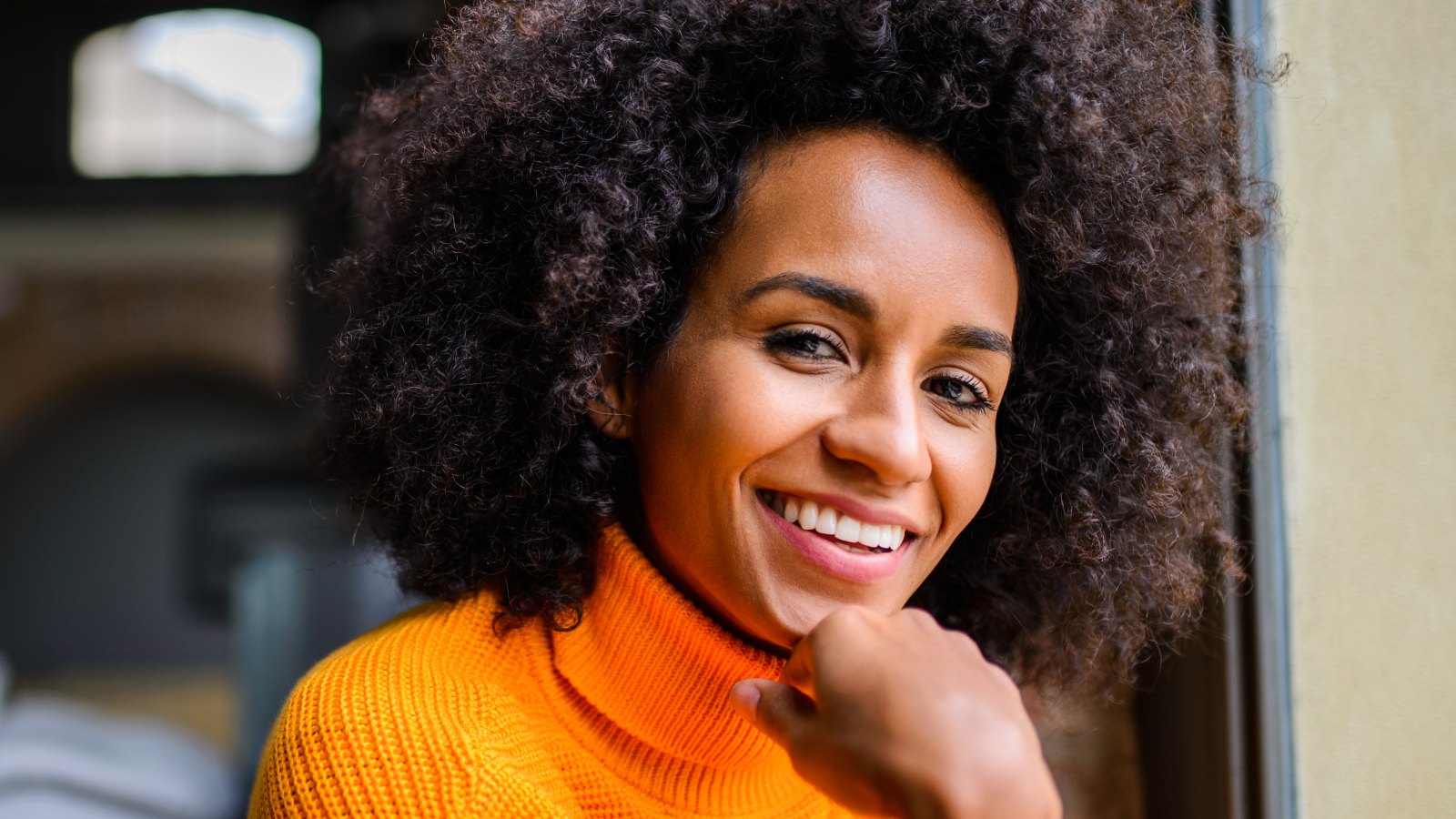 woman with big curly hair smiling while sitting down wearing a bright orange sweater