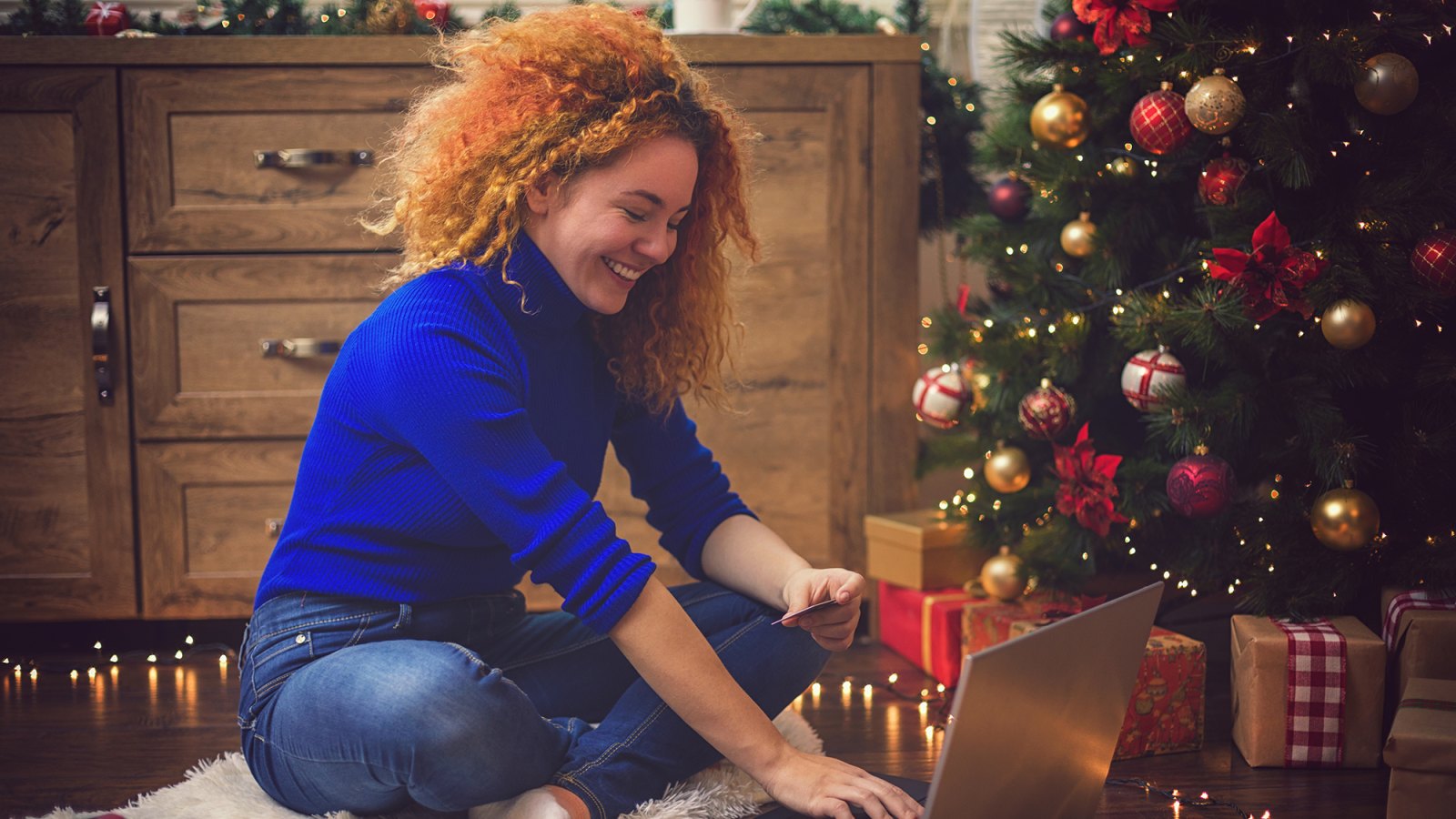 Young beautiful girl with red curly hair ,enjoying at home in a cozy Christmas atmosphere while using lap top for online shopping.