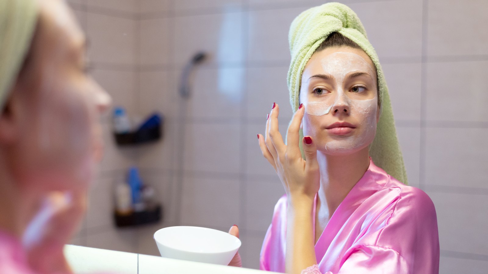 Beautiful young woman putting facial mask on her face in front of the mirror at home.