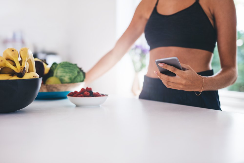 Cropped shot of young woman with mobile phone in kitchen. Fruits and vegetable on kitchen counter.