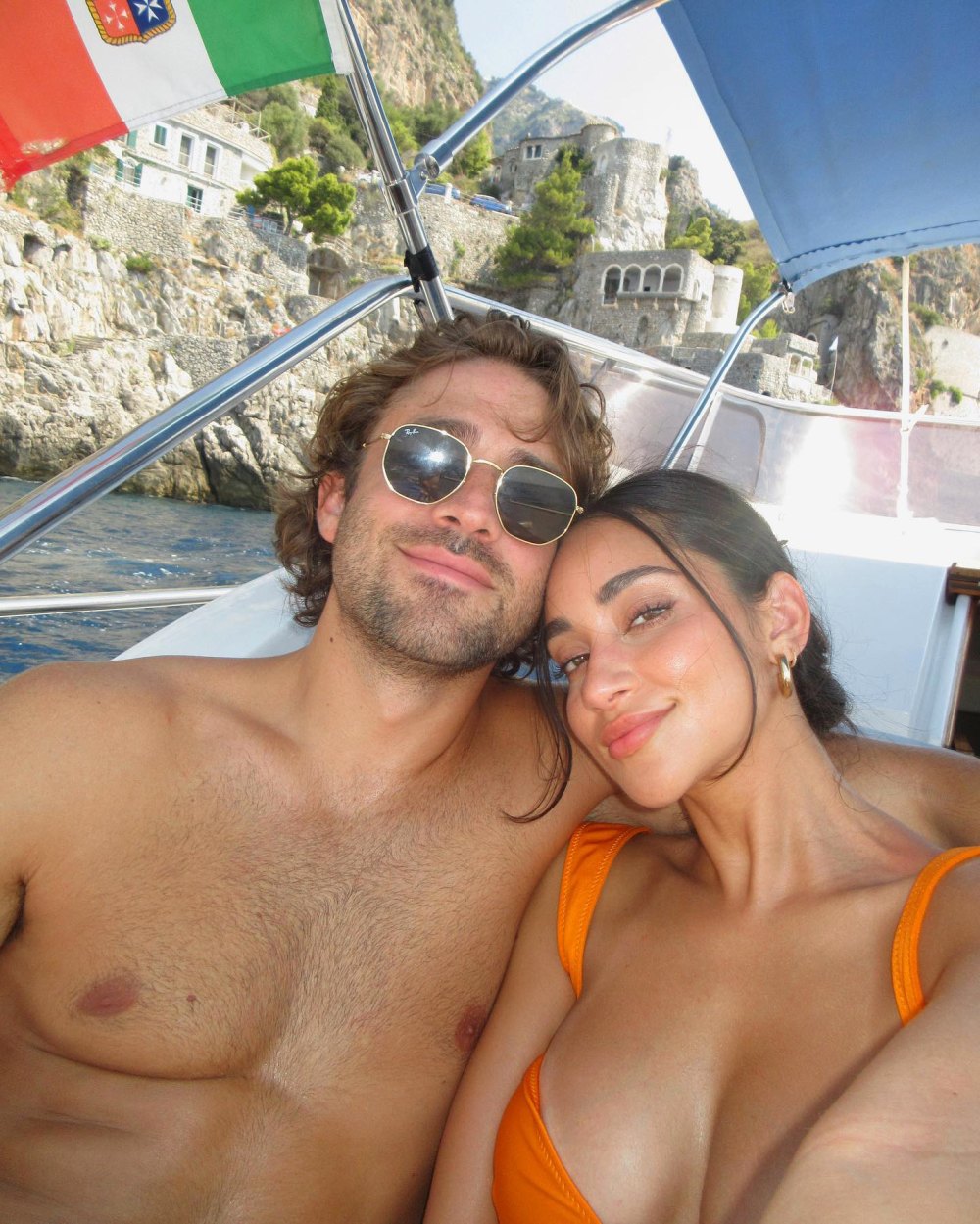 Greg Grippo shirtless, wearing sunglasses with arm around Victoria Fuller wearing orange bikini as they smile on a boat on the Amalfi Coast