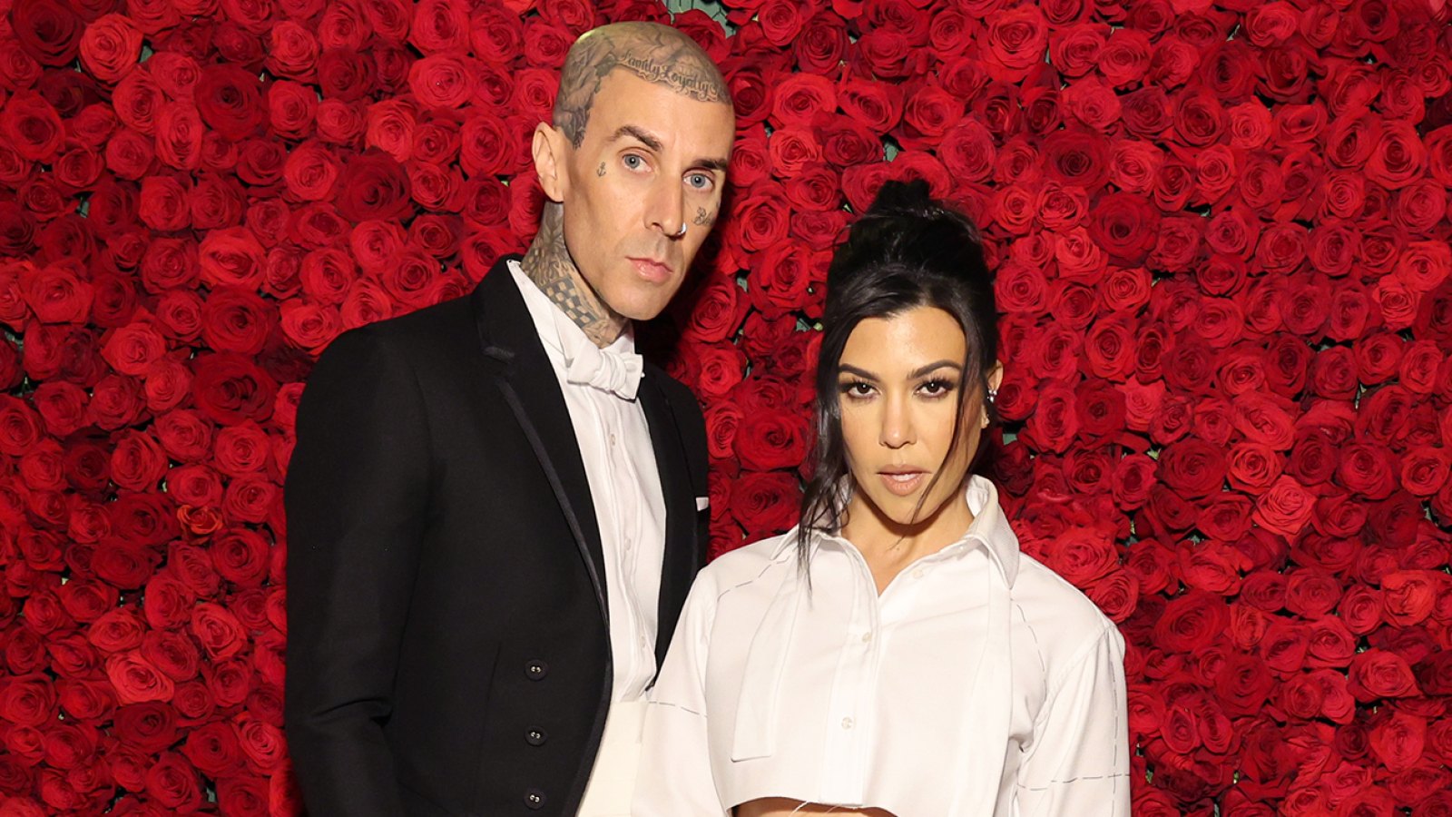Travis Barker and Kourtney Kardashian hold hands and pose in front of a wall of red roses at the Met Gala