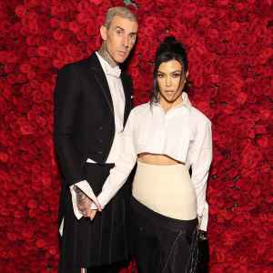 Travis Barker and Kourtney Kardashian hold hands and pose in front of a wall of red roses at the Met Gala