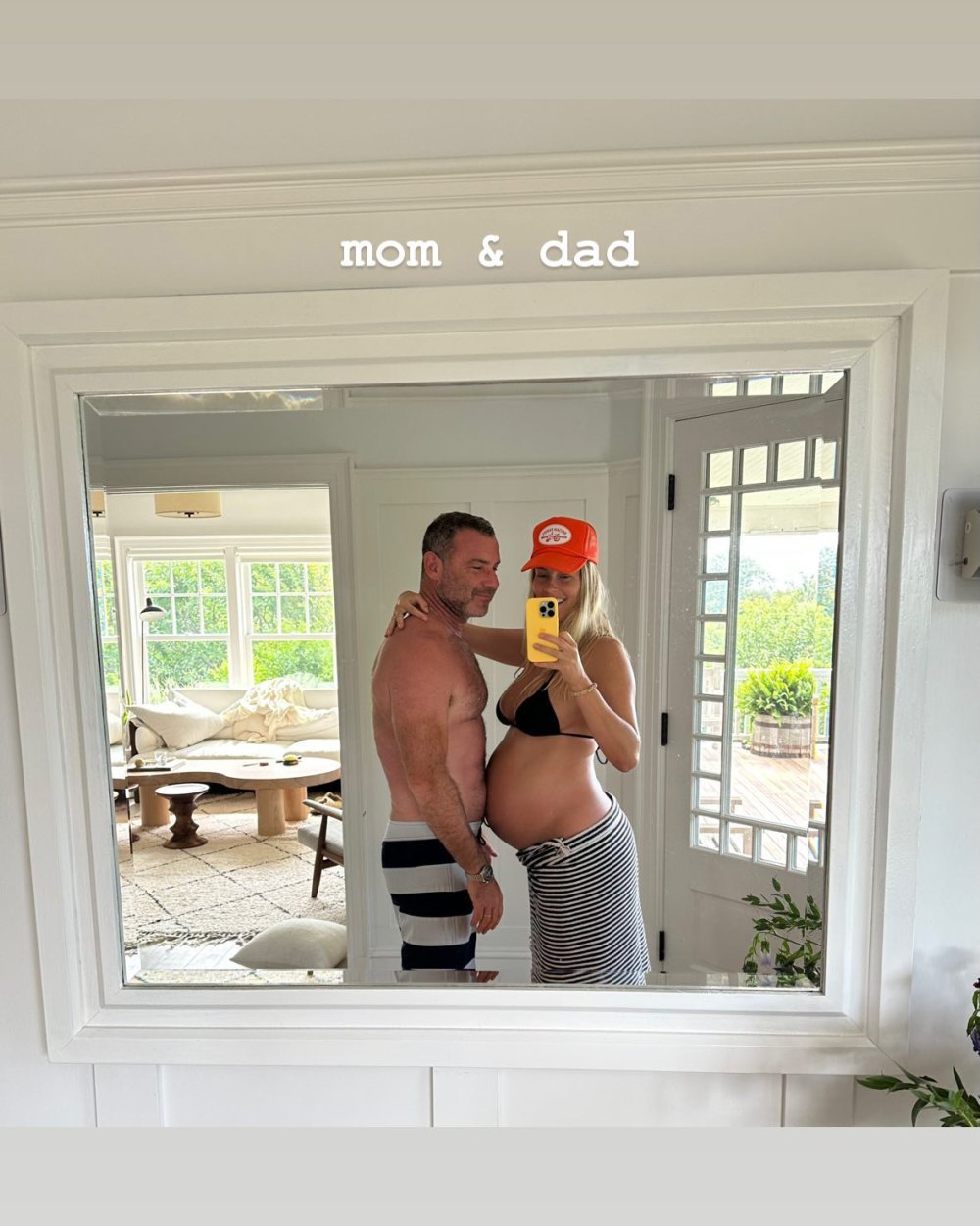 Liev Schreiber Shirtless in Striped Board Shorts Smiles in Mirror Selfie with Pregnant Taylor Neisen wearing a Black Bikini top, Orange Hat, and Striped Skirt holding Yellow Phone