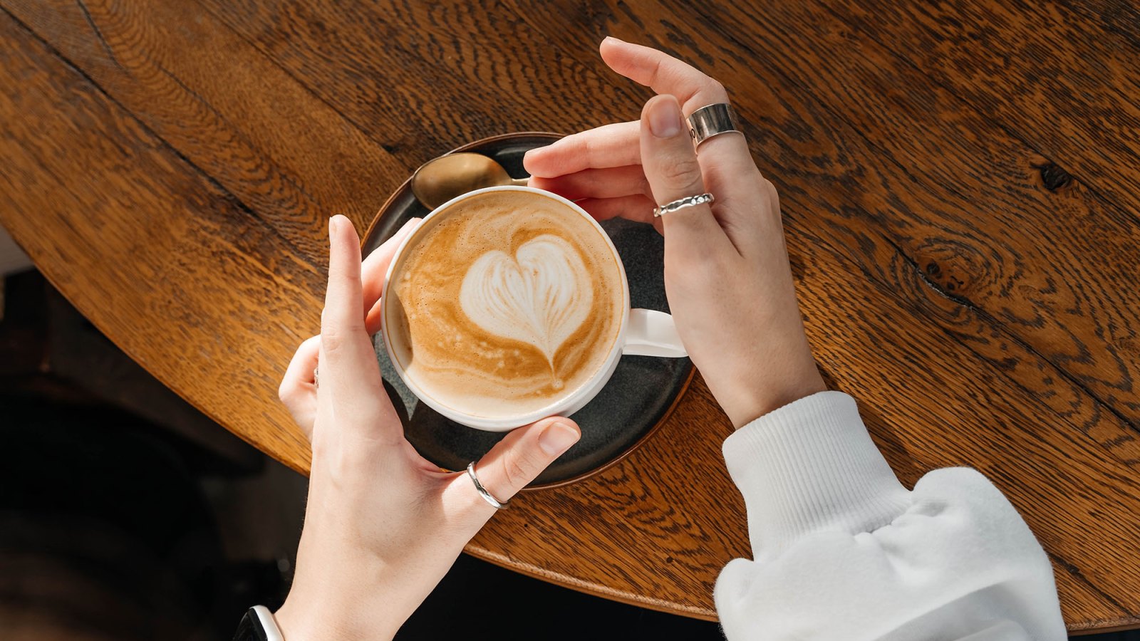 Cappuccino in a white ceramic cup with women's hand on a wooden table. Top view.