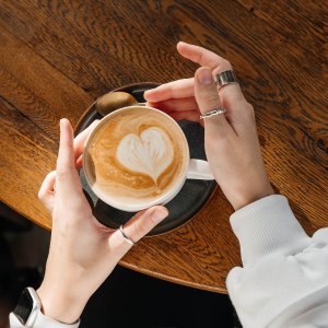 Cappuccino in a white ceramic cup with women's hand on a wooden table. Top view.