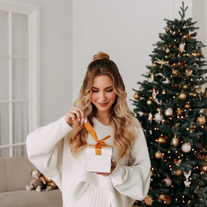 The concept of Christmas. Portrait of a smiling young woman with curly hair in a knitted sweater holding and opening a Christmas gift on the background of a Christmas tree in a bright interior on a holiday at home