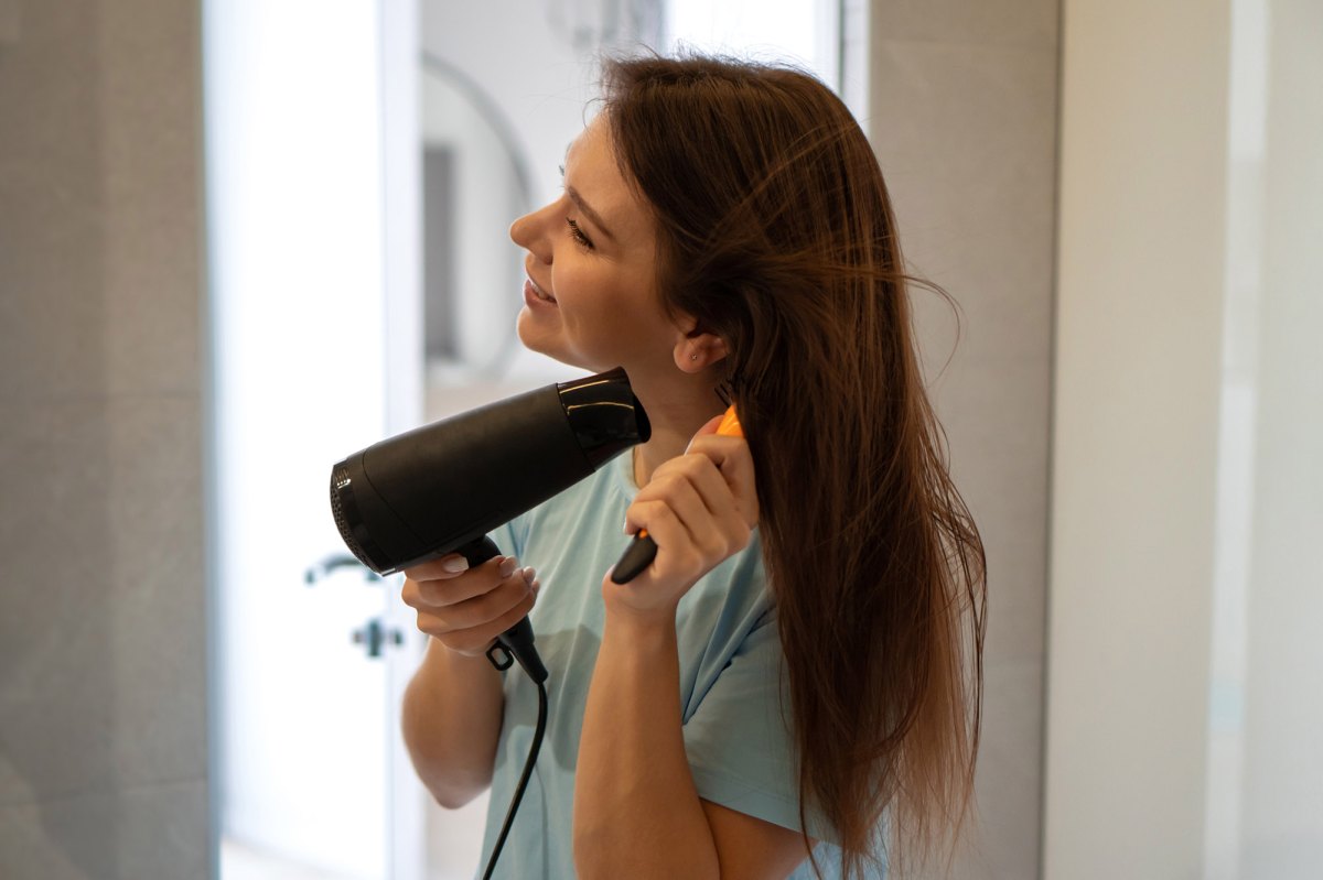 Young woman dries her long brown hair with a hair dryer looking to the mirror. Attractive female profile view. Self care, body care, wellness concept