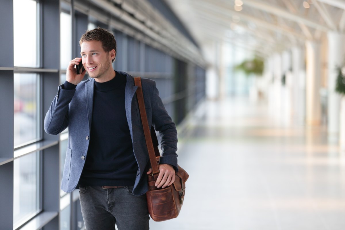 highly rated messenger bag Urban business man talking on smart phone traveling walking inside in airport. Casual young businessman wearing suit jacket and shoulder bag. Handsome male model in his 20s.