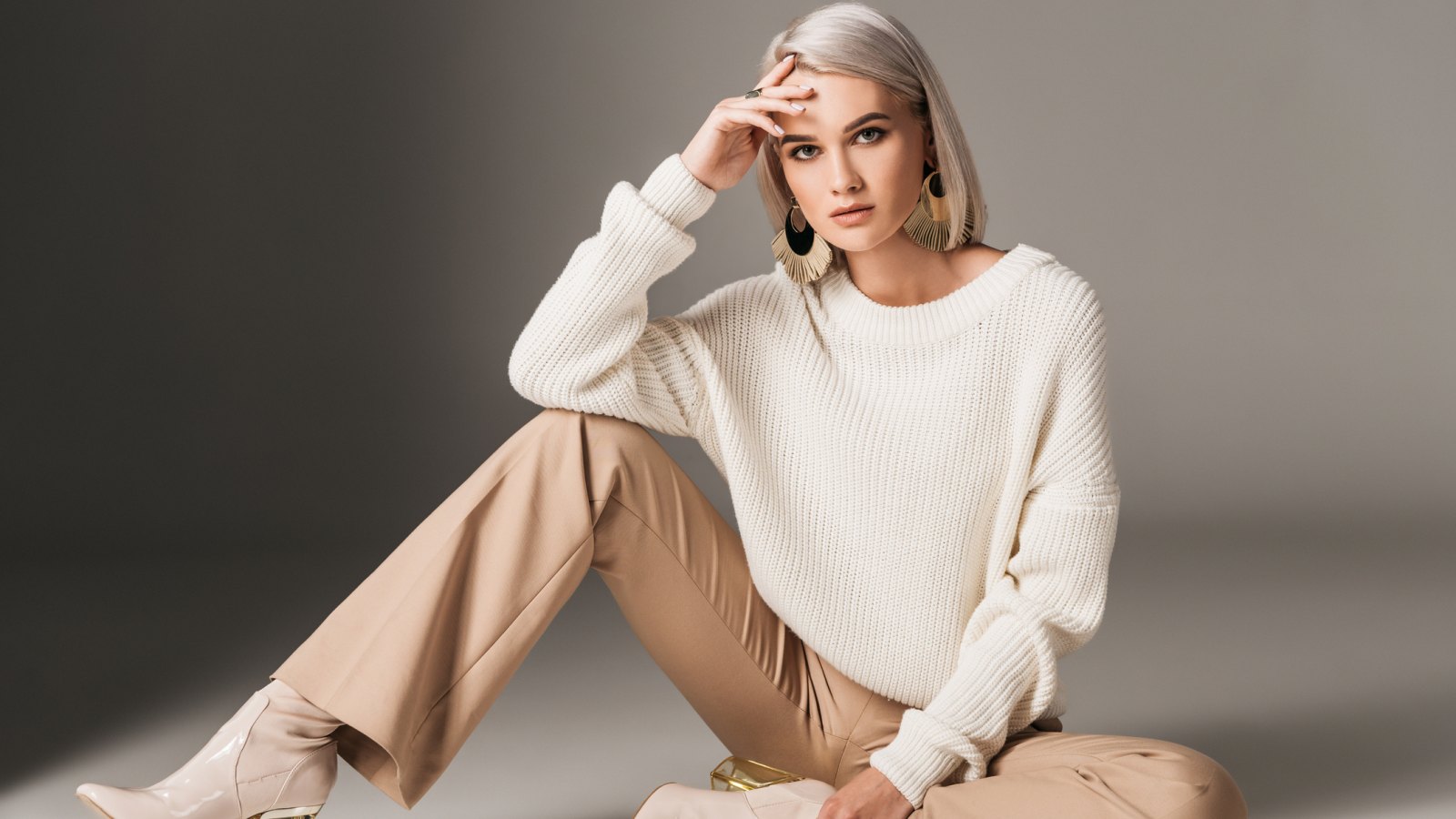 attractive fashionable woman posing in white trendy sweater, beige pants and autumn heels, on grey
