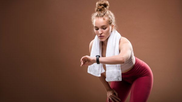 Tired young blond sportswoman in activewear looking at fitbit on her wrist while doing difficult physical exercise against brown background