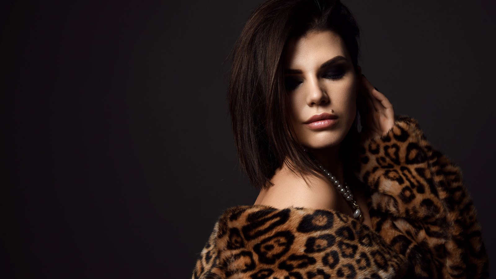 Portrait of young pretty brunette woman model in leopard fur coat with naked shoulders in pearl necklace looking down