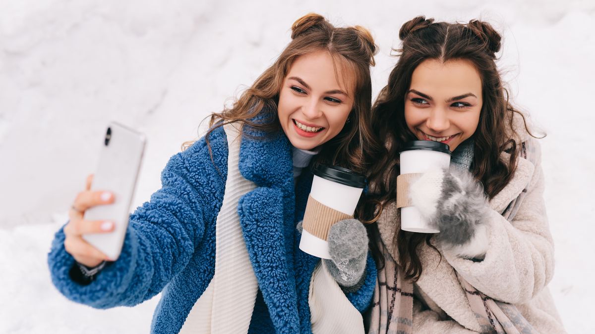 Two happy girls millennials teenagers friends are walking, laughing, taking selfies using wireless technology and a mobile phone, skating on a skating rink in a winter forest during the Christmas holidays. A couple of women in love spends time together in nature