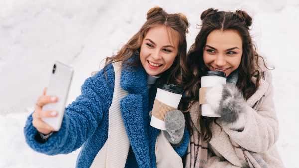 Two happy girls millennials teenagers friends are walking, laughing, taking selfies using wireless technology and a mobile phone, skating on a skating rink in a winter forest during the Christmas holidays. A couple of women in love spends time together in nature