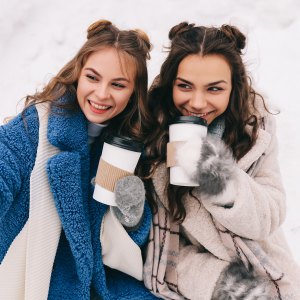 Two happy girls millennials teenagers friends are walking, laughing, taking selfies using wireless technology and a mobile phone, skating on a skating rink in a winter forest during the Christmas holidays. A couple of women in love spends time together in nature