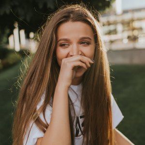 Stylish young blonde with extra long hair laughs cheerfully and looks away. She covers her mouth with palm. Sun illuminates her gently, well-groomed park is on the background.