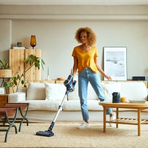 Smiling young woman cleaning carpet with vacuum cleaner at home. Female is doing housework in living room. She is standing against sofa.