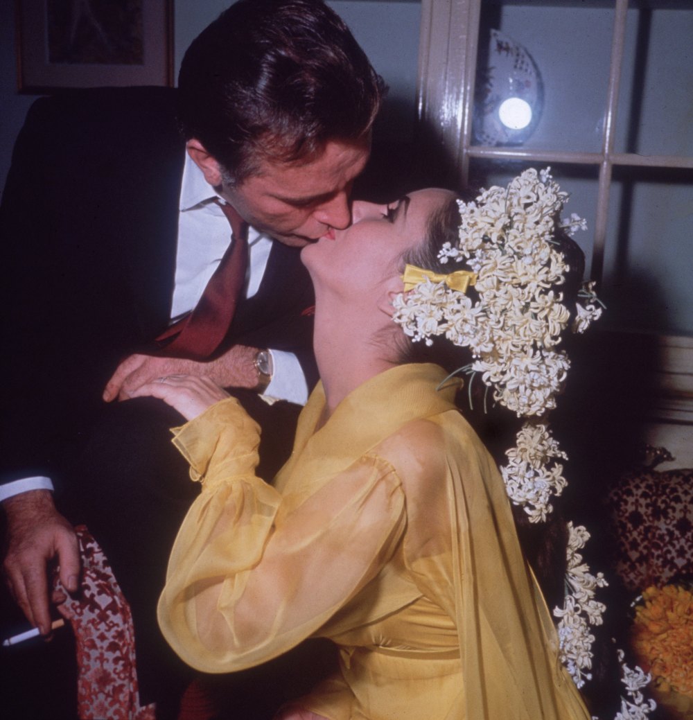 15th March 1964: British-born Elizabeth Taylor sits on a sofa kissing her fifth husband, Welsh actor Richard Burton, while he leans over her on their first wedding day. Taylor is wearing a yellow gown and has flowers in her hair. (Photo by Hulton Archive/Getty Images)