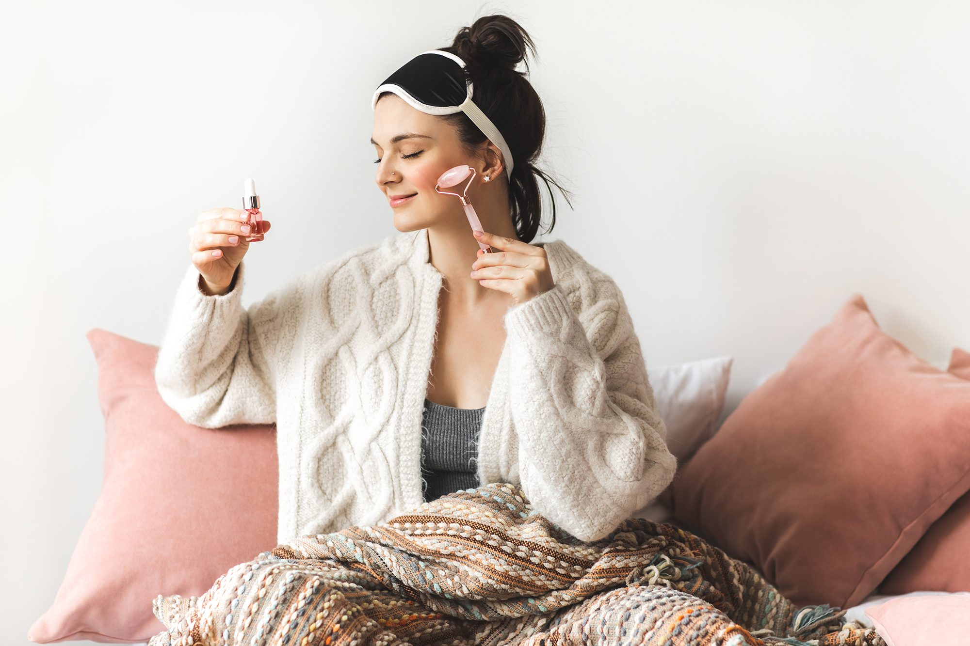 A girl with nice skin and her hair in a bun is sitting down comfortably leaning on pillows, legs covered with blanket. She is wearing a soft white fluffy cardigan and a facemask on her head. Beautician is holding up a small bottle of skincare oil and applying it as she's massaging her face with a special roller. It's very relaxing and she is smiling pleasantly. Self care concept.