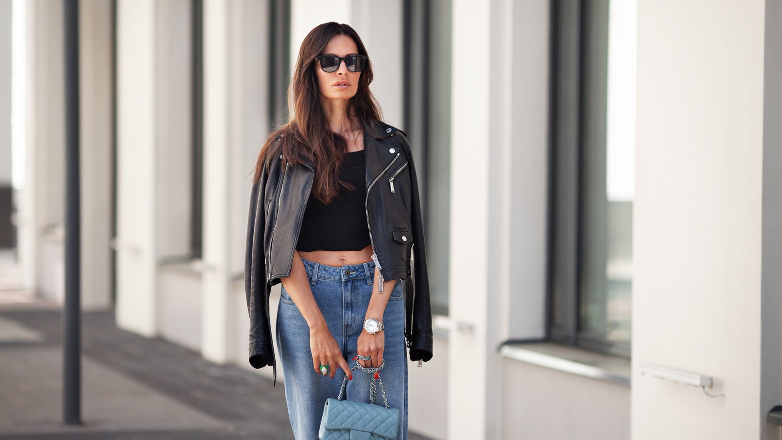 Portrait of gorgeous brunette woman standing city street. Fashion model wears black leather jacket, jeans, short top, small handbag on chain. Street style.