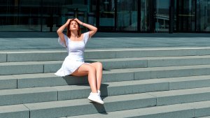 Beautiful young woman sitting on stairs and enjoys the bright sunlight on a summer day. Young woman wearing a white dress and sneakers sitting on stairs in front of a hotel