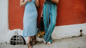 Conceptual and bright image of two females standing against a red wall in the streets of Positano. They are wearing fashionable and light clothing, appropriate for warm weather and comfortable walking sandals, perfect for exploring the winding streets of Italy.