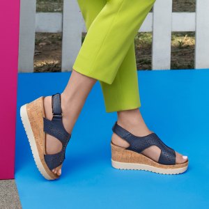 Outdoor scene featuring a woman modeling stylish navy blue wedge sandals, cork soles and perforated straps. stands on a bright blue floor, pink wall, wearing lime cropped pants, greenery background.