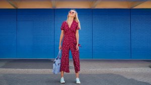 Pretty blond woman wearing burgundy jumpsuit and sunglasses while walking against the blue wall and holding cerulean tote bag