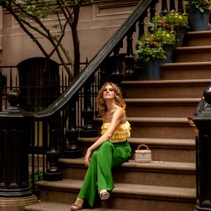 Red-haired young woman in stylish bright yellow top and green trousers elegant sandals sitting on the potted flowers porch stairs on Manhattan West village.