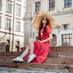 Summer vibes. Retro fashion: straw hat, red polka-dot dress and white sneakers. Fashionable girl sitting on the stairs of the town square.
