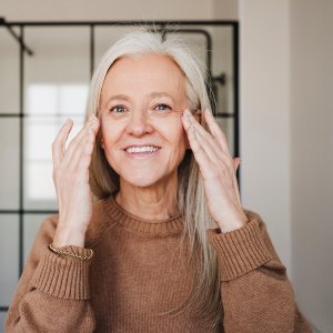 Happy mature woman applying moisturizer on face at home