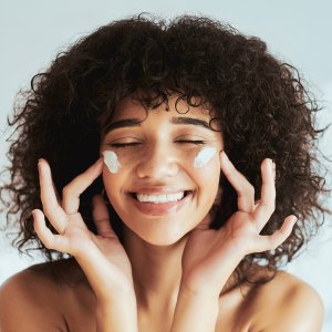 Moisturizer, cream on face in cosmetics, beauty or skincare promotion on white wall background. Facial, happy person or model smile with lotion, essential oil or sunscreen application
