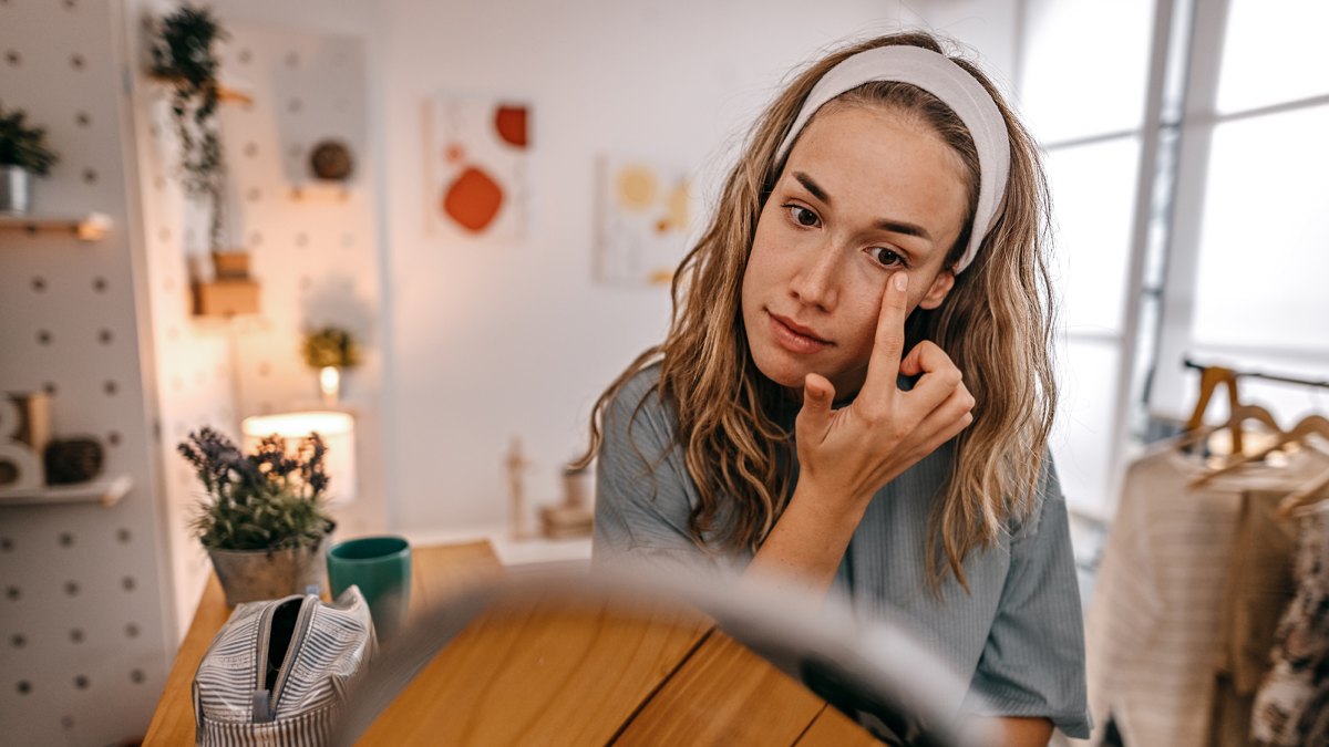 Young beautiful woman looking in mirror tightening her under eye skin with finger at home