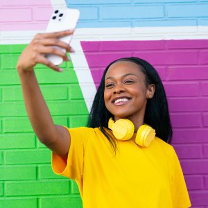 Brightly dressed woman with yellow headphones enjoys taking a selfie, with a vibrant urban wall behind.
