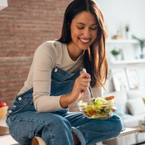 Shot of smiling woman eating healthy salad while sitting on the kitchen table at home.