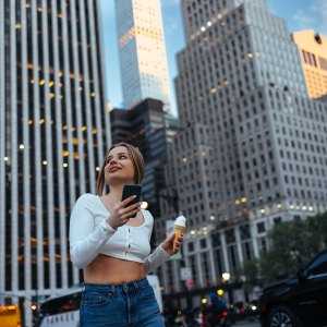 A woman gazes upward while holding her phone and ice cream cone, city buildings behind her