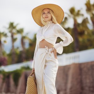 Stunning blonde woman in summer beach outfit relaxing outdoors against sea resort and palm trees on the background. A fashionable romantic young adult lady wearing a trendy vintage straw hat, white blouse, and skirt, standing at the sea coast