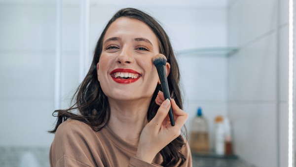 A young woman wearing red lipstick is looking at the camera with a big smile while applying blush to her cheeks.
